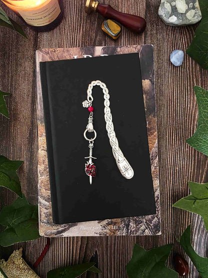 Marque-page "Le voeu de l'épée", un coeur humain transpercé par une épée, avec une breloque main au dessus qui tient l'épée ainsi qu'une pierre naturelle de corail rouge. Le signet est en métal et évoque un bijou de livre très beau pour les romans de fantasy, dark fantasy et romantasy